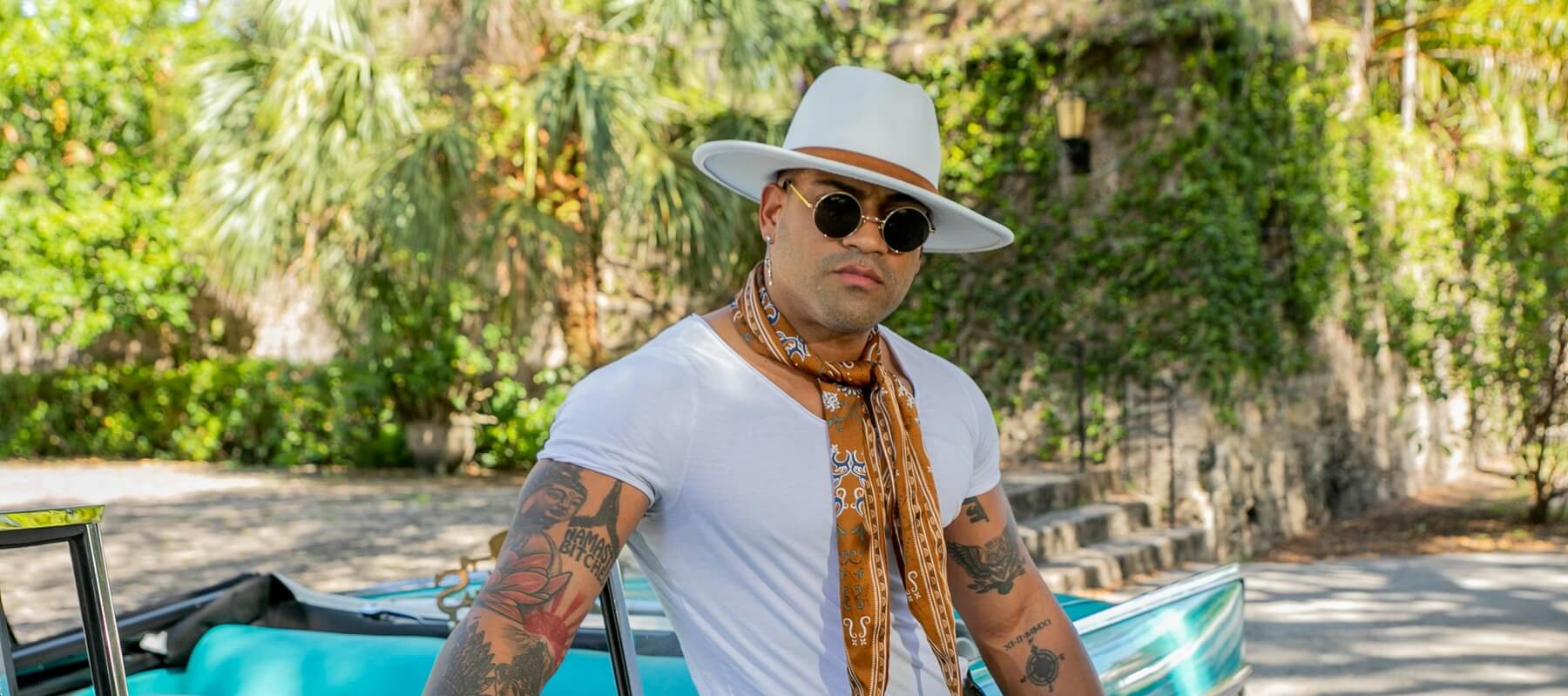 Man wearing a white hat, sunglasses, and a scarf standing next to a vintage car with greenery in the background